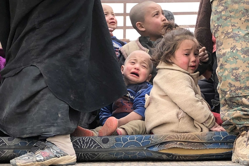 A group of children sit on the ground crying between the feet of two men, one in army fatigues.