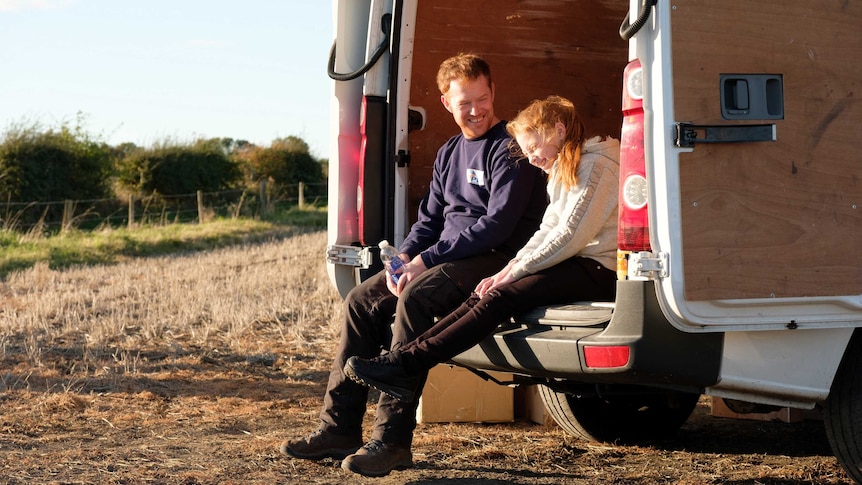 A man and girl sit in the back of a van parked in a paddock