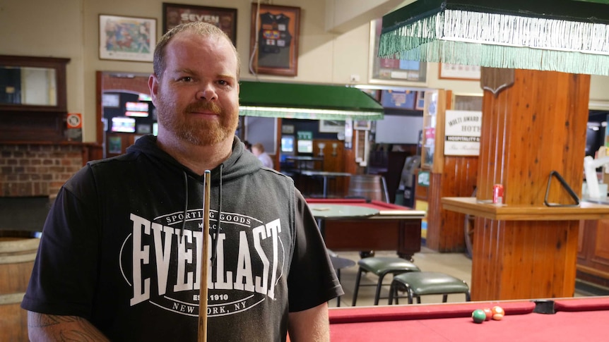 A man stands in front of a pool table in a pub, holding a pool cue in front of him.