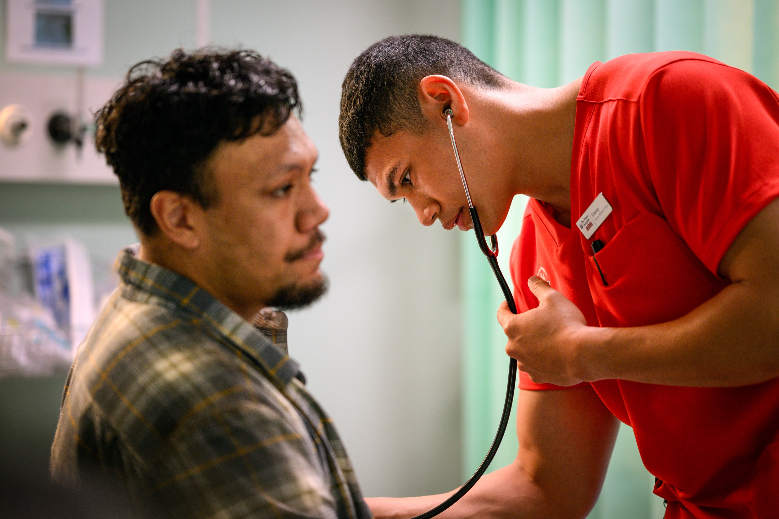 A TV still of Ioane Sa'ula, mid-20s, Samoan Australian, in orange scrubs, holding a stethoscope to Aliki Matangi's chest.