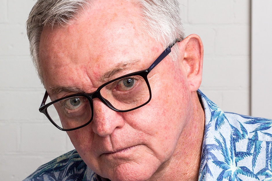 An older man wearing glass in front of a white wall.