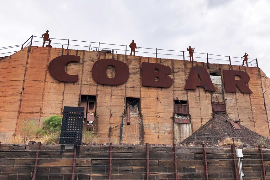 Large lettering of Cobar on a concrete wall, with steel silhouettes of miners carrying picks