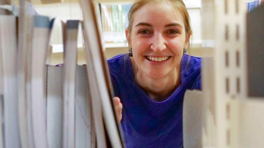 A young fair woman with blonde hair smiles into the camera, she is wearing a royal blue shirt and peers through library shelves.