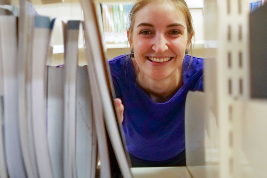 A young fair woman with blonde hair smiles into the camera, she is wearing a royal blue shirt and peers through library shelves.