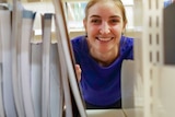 A young fair woman with blonde hair smiles into the camera, she is wearing a royal blue shirt and peers through library shelves.