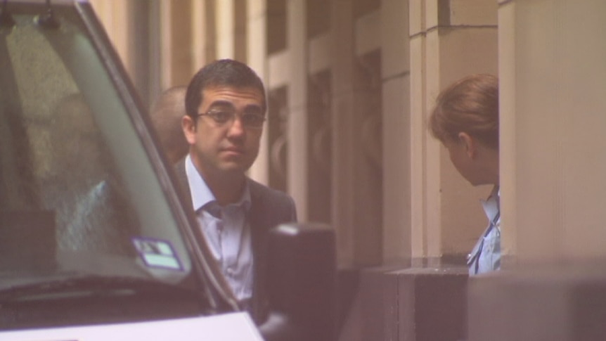 A man in glasses is led by prison staff from a van into a court building.