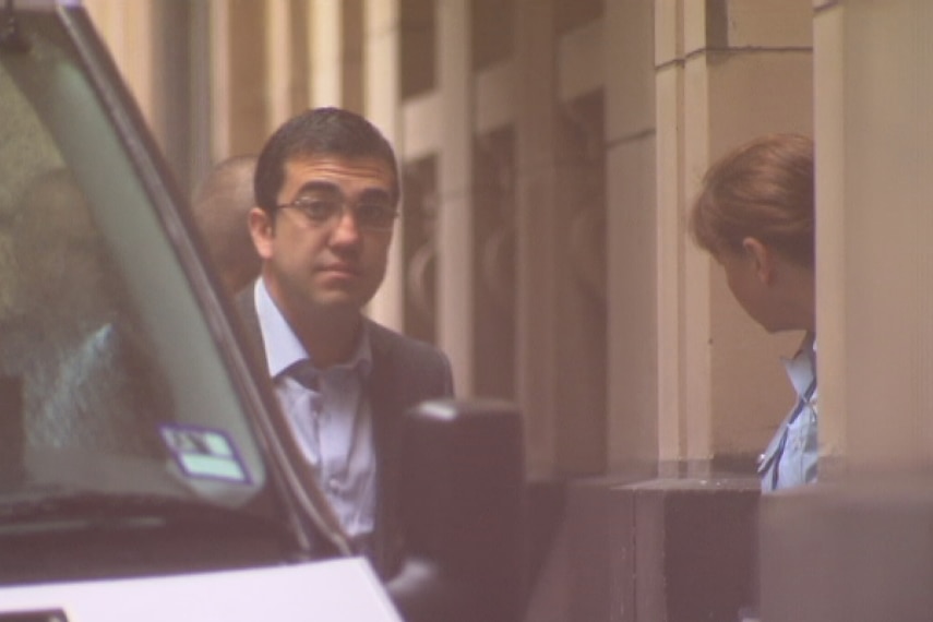 A man in glasses is led by prison staff from a van into a court building.