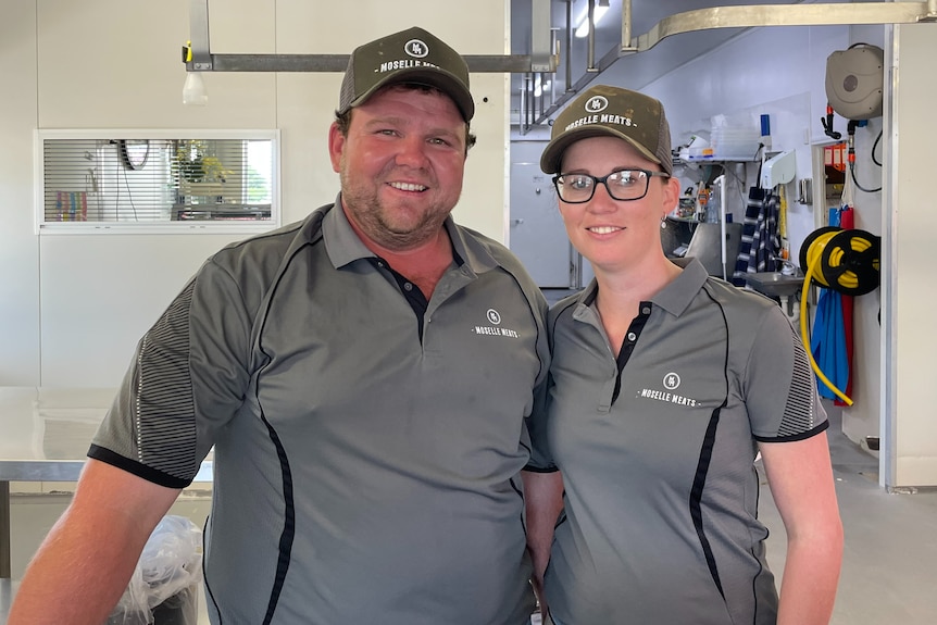 Man and woman with matching grey shirts and hats on stand next to eachother in butcher smiling