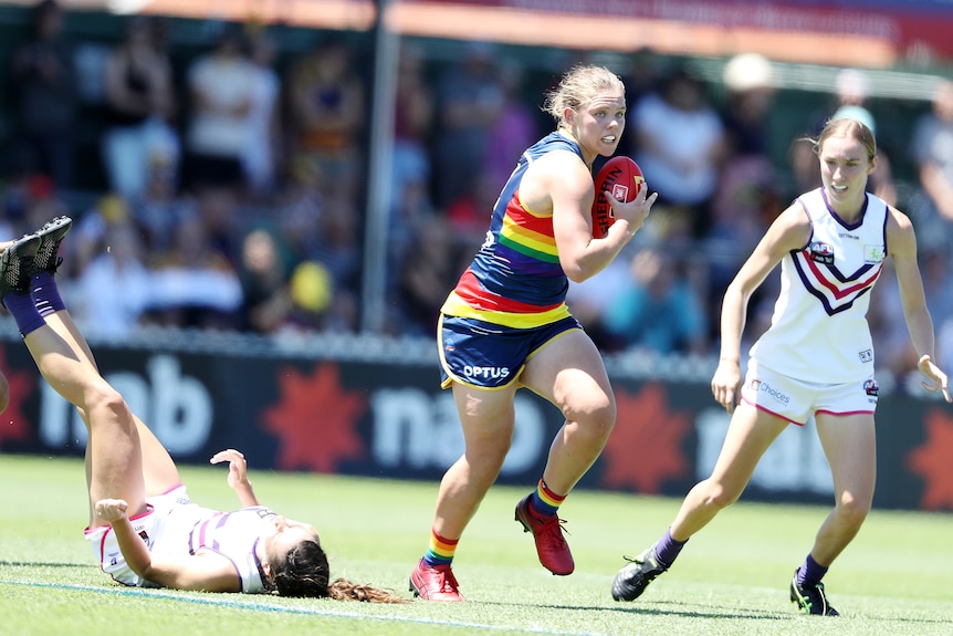 An AFLW player runs downfield with a defender next to her and another lying on the ground behind her.