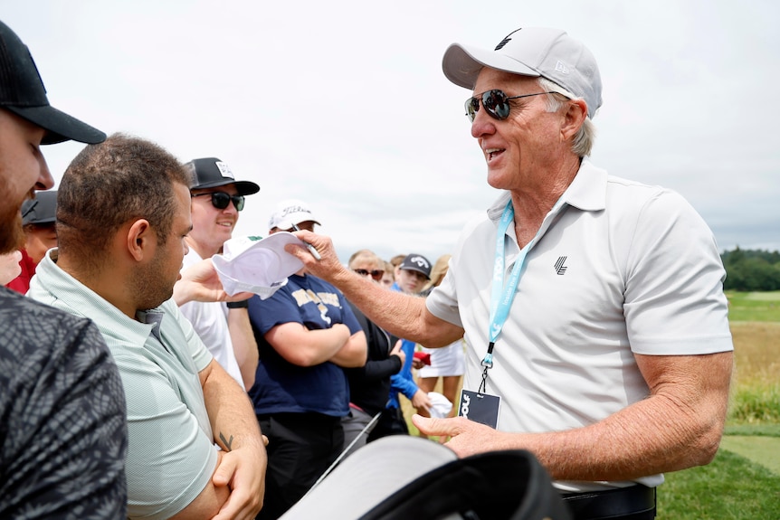 A fit older white man with dark glasses and white hair smiles as he signs autographs for fans on a golf course.