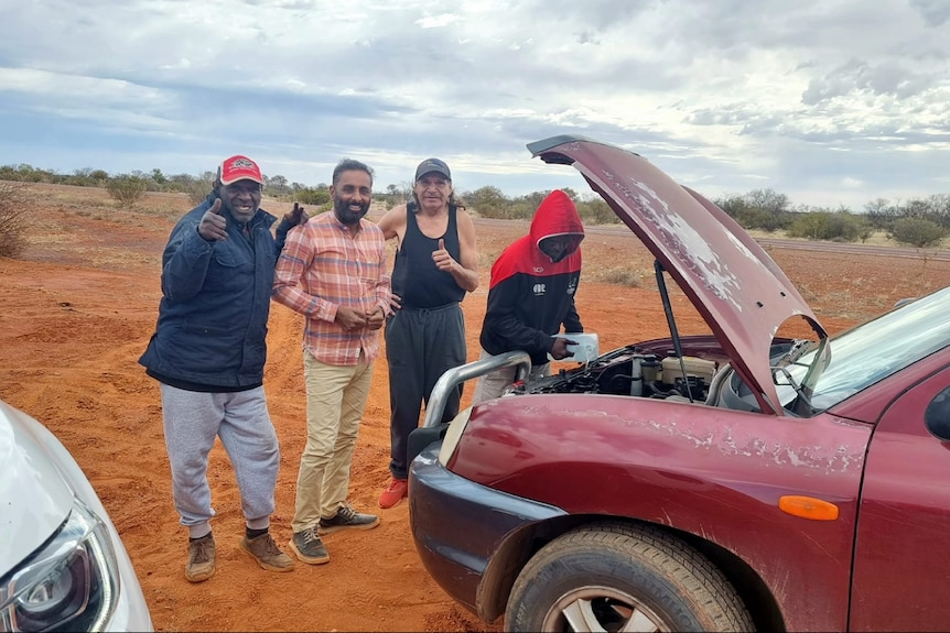 Mintu does a thumbs up gesture with some First Nations people in Coober Pedy, one guy is repairing a car 