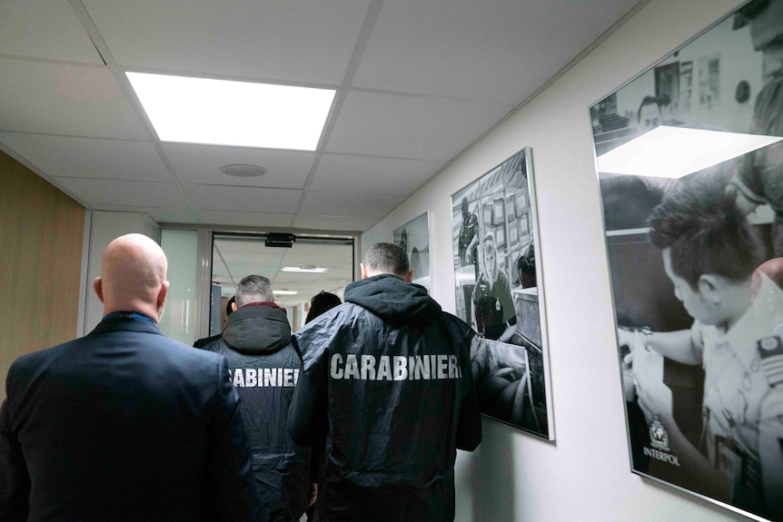 People in black jackets with Carabinieri written on the back walk through a hall lined with black and white photos. 