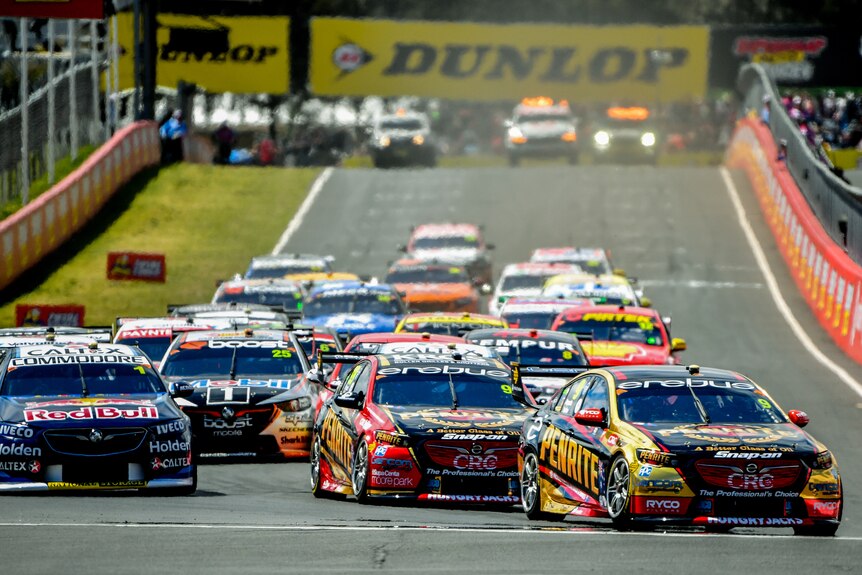 A group of cars race down a straight, with the leaders about to turn