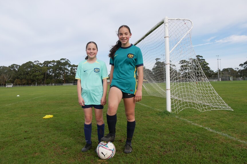 Two girls wearing Australian soccer shirts standing behind a ball. 