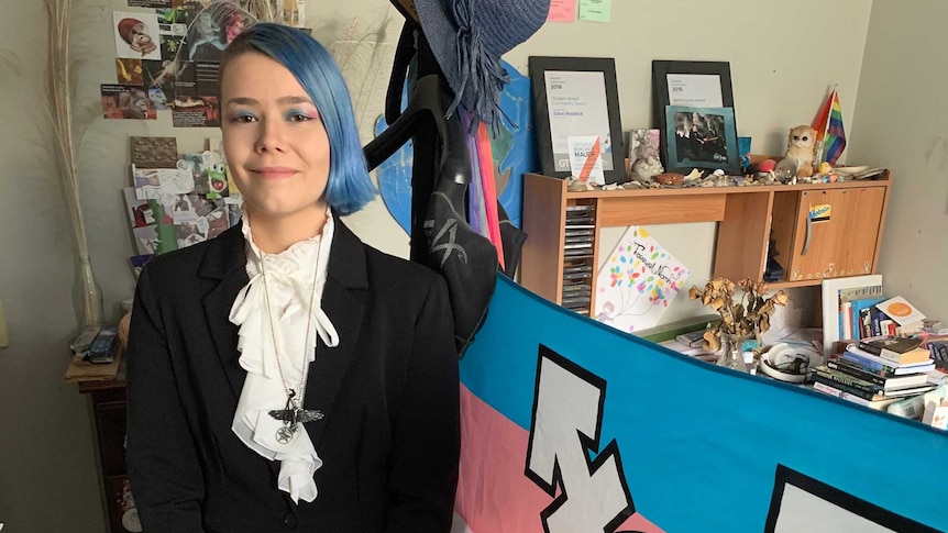 Eden Meddick stands in his bedroom in front of the pink and blue transgender flag.