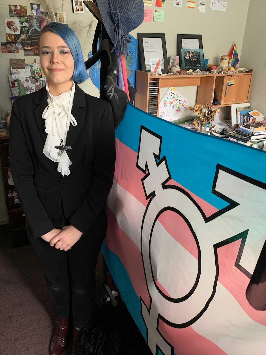 Eden Meddick stands in his bedroom in front of the pink and blue transgender flag.