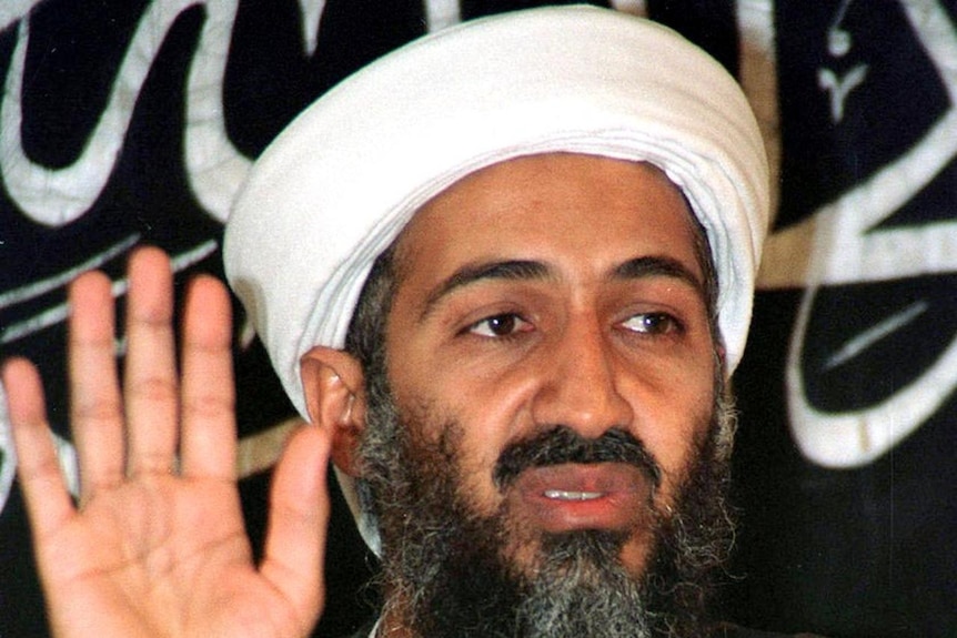 A turbaned Osama bin Laden, with his right hand raised, is sitting in front of a black and white Al Qaeda flag