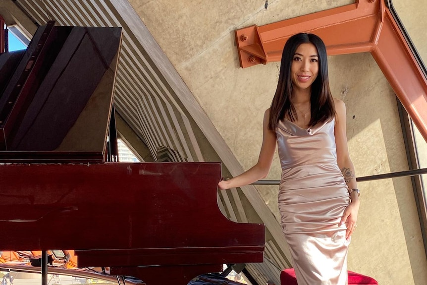 A young woman wearing a pink dress smiling with her hand on a grand piano.