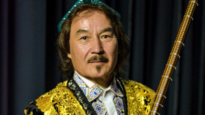 Uyghur musician Shohrat Tursun stands holding his lute. He is wearing a black jacket with gold embroidery.