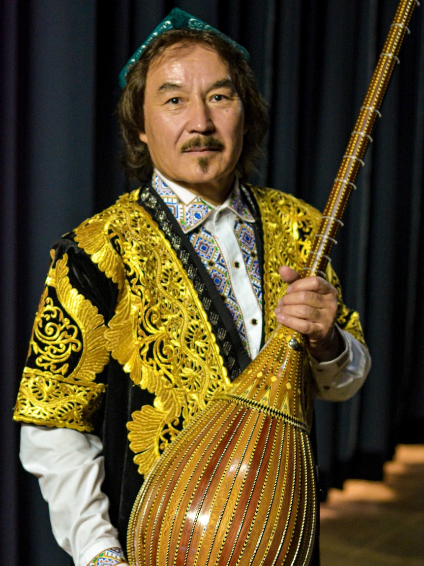Uyghur musician Shohrat Tursun stands holding his lute. He is wearing a black jacket with gold embroidery.