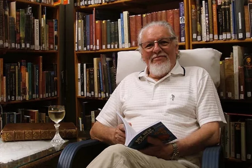 An older man with grey hair and goatee sitting in a liberary with a glass of white wine next to him, reading a book.