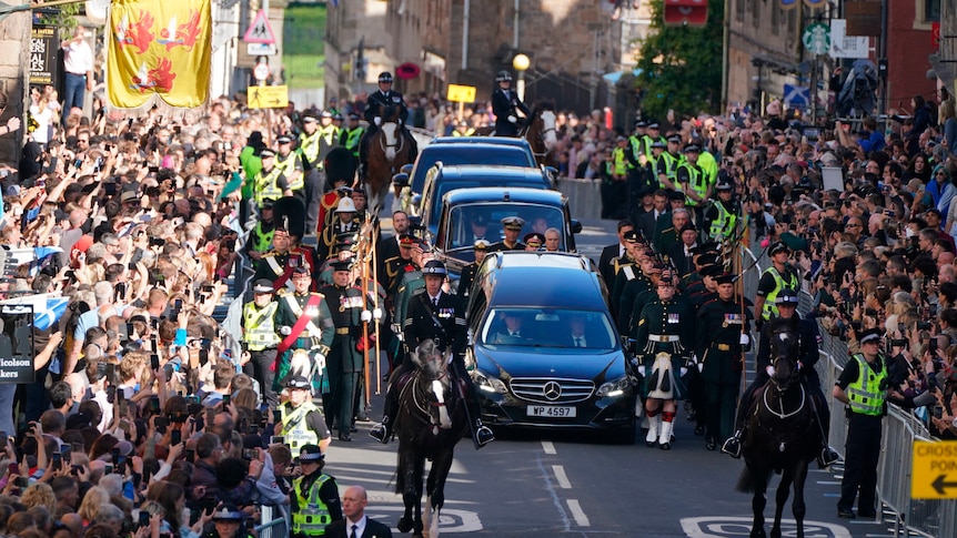 Crowds of people look on as two horses lead the procession of cars and the Queen's coffin down a street. 