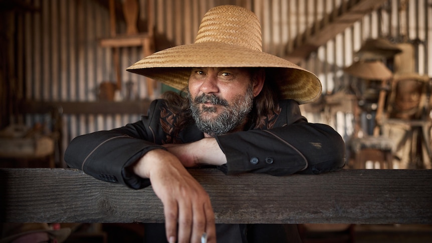 A bearded Indigenous man in a wide-brimmed straw hat smiles for a photo inside a tin shed.