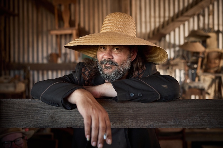 A bearded Indigenous man in a wide-brimmed straw hat smiles for a photo inside a tin shed.