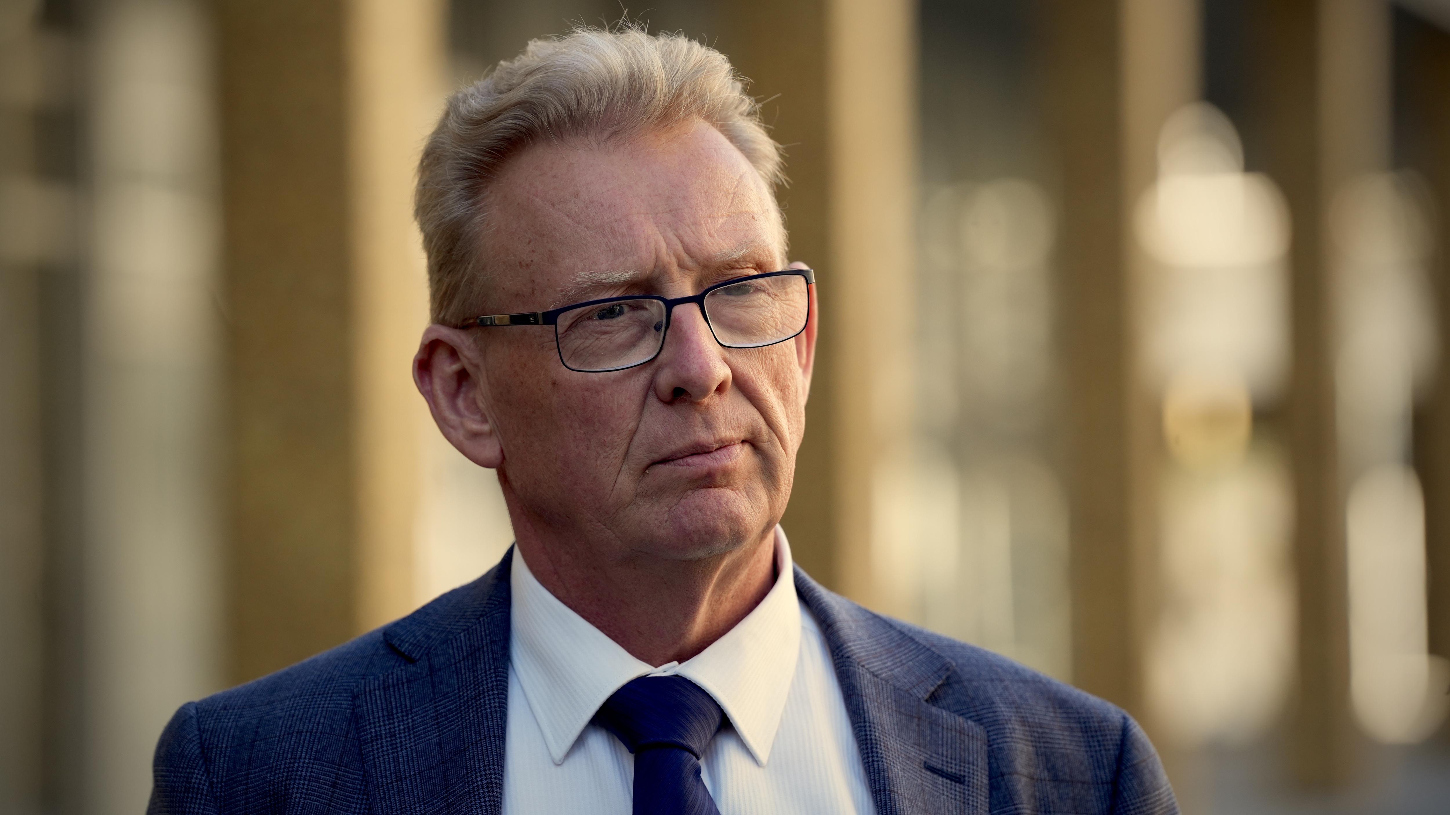 A man stands outside listening, he wears a suit and tie and glasses and has blonde hair.