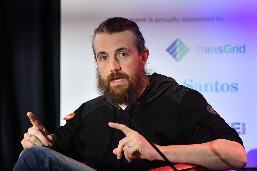 A man with a bushy beard and hair tied back sits at a table on a stage, talking with a projector screen behind him.