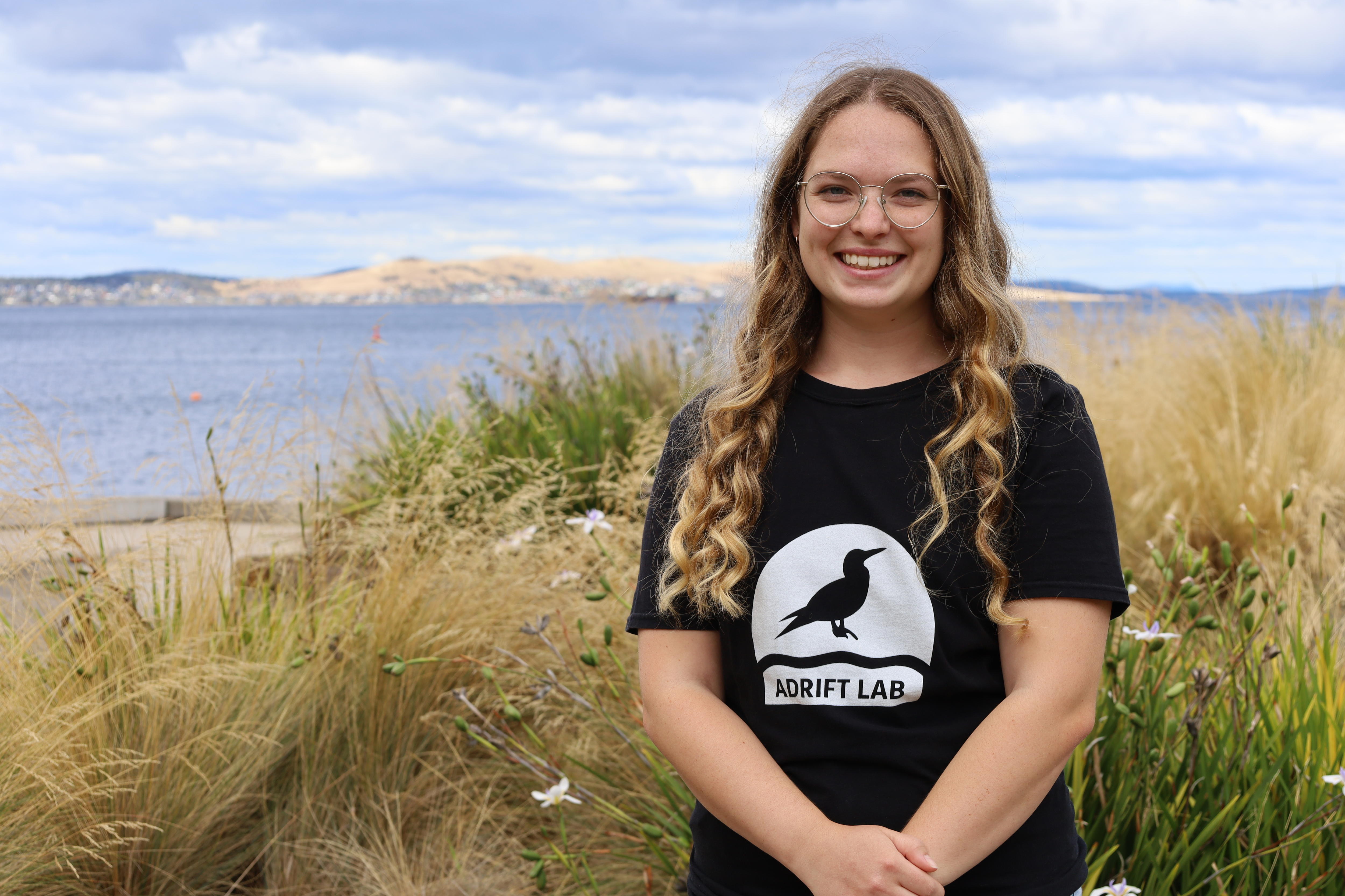 Person standing smiling on beach. Black tshirt and blonde hair.