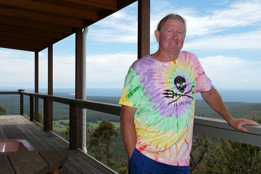 A man with grey hair and a tie-dyed t-shirt stands on his balcony.