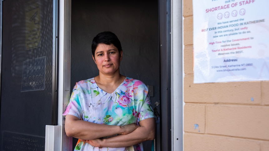 A woman stands in a doorway with her arms folded with a sign in the periphery that says the business is closing. 