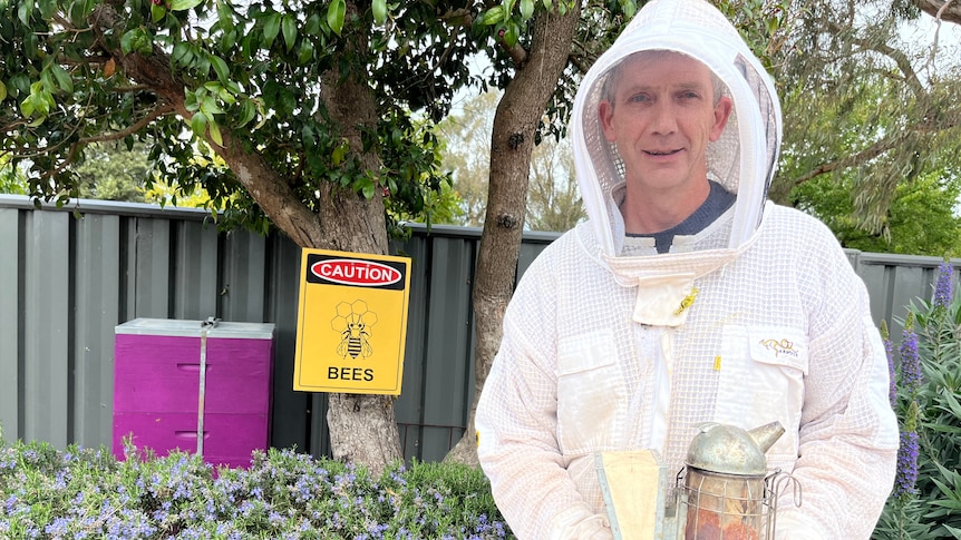 A man in protective clothing holds bee handling equipment in front of a sign reading 'caution: bees'.