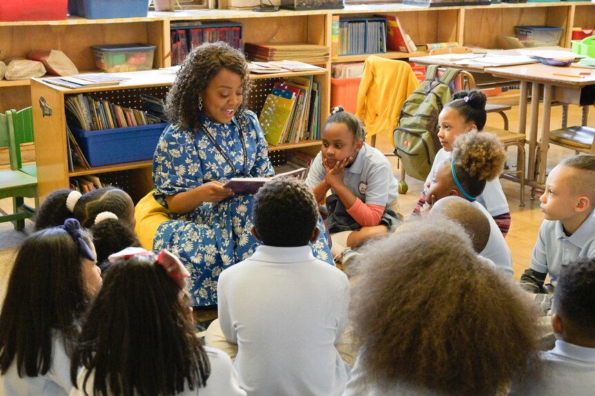 A black woman with curly dark hair wears a blue and white floral dress and sits in front of a class of children reading a book.