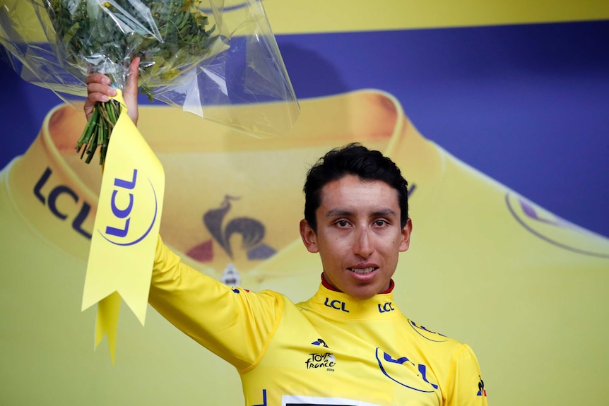 Egan Arley Bernal Gomez wearing a yellow jersey holds up a bouquet of flowers in the air