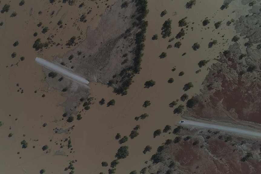 An aerial view of a road submerged in brown floodwater with a car at the edge of the water.