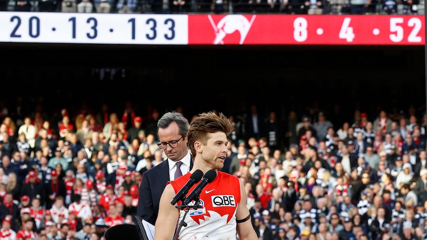 A Sydney Swans captain stands at a microphone while behind him the scoreboard says 20.13.133 for Geelong and 8.4.52 for Sydney.