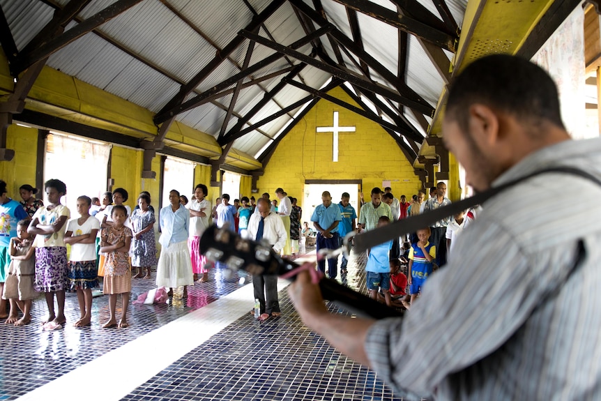 Un evento de la iglesia en PNG.  Se ve a los fieles de pie mirando a un hombre tocando su guitarra.