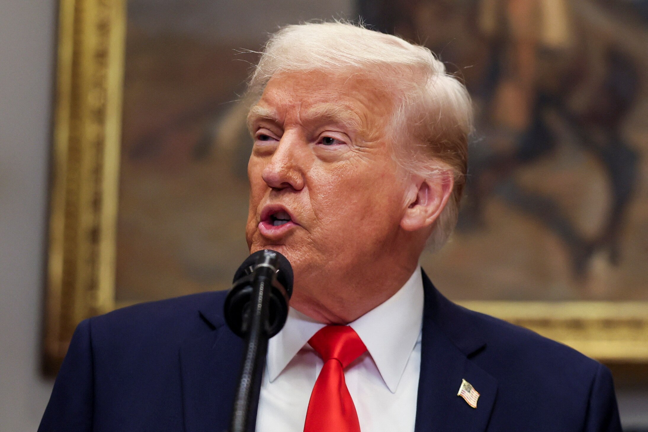 Donald Trump with lips pursed mid-speech wearing a dark suit jacket and red tie in front of a portrait.