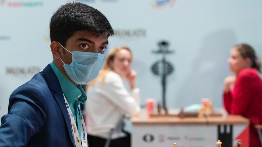 A man sits at a chess board wearing a face mask. Two people are playing chess behind him.