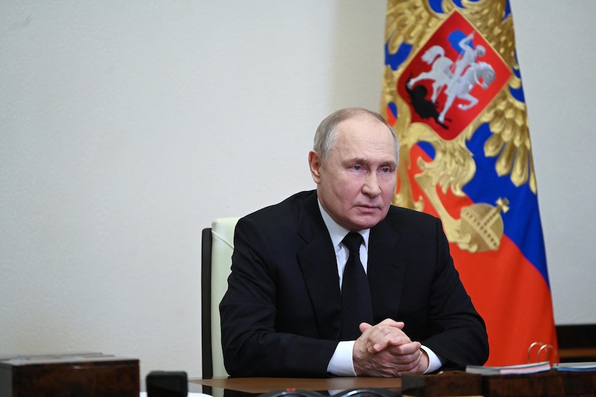 Russian President Vladimir Putin sits as a desk with a Russian flag behind him.