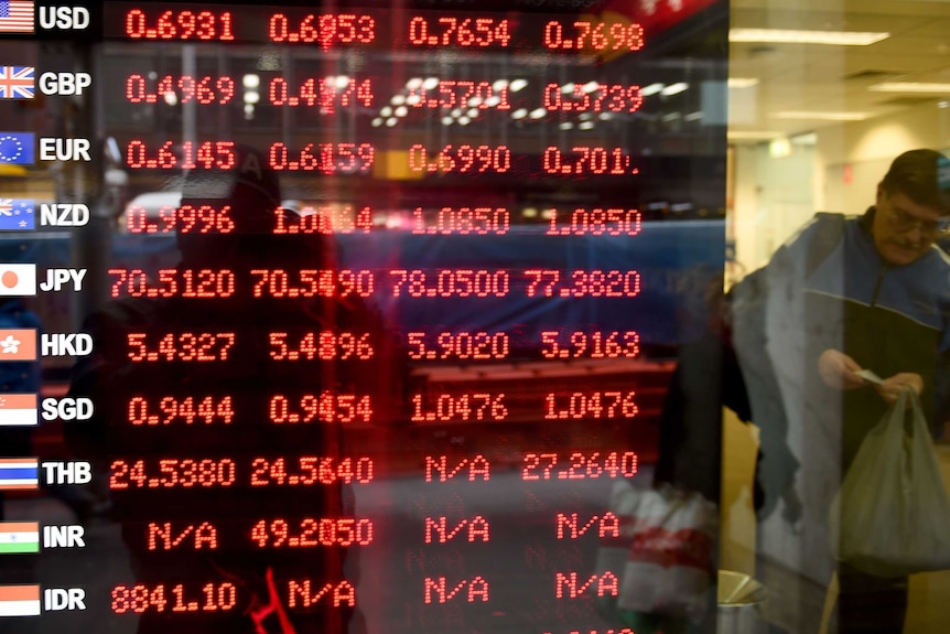 A man leaves a bank next to an electronic screen displaying currency rates in red.