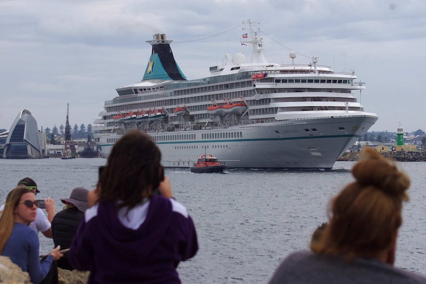 The Artania cruise ship pulling away from Fremantle Port with people in the foreground taking pictures on their phones.