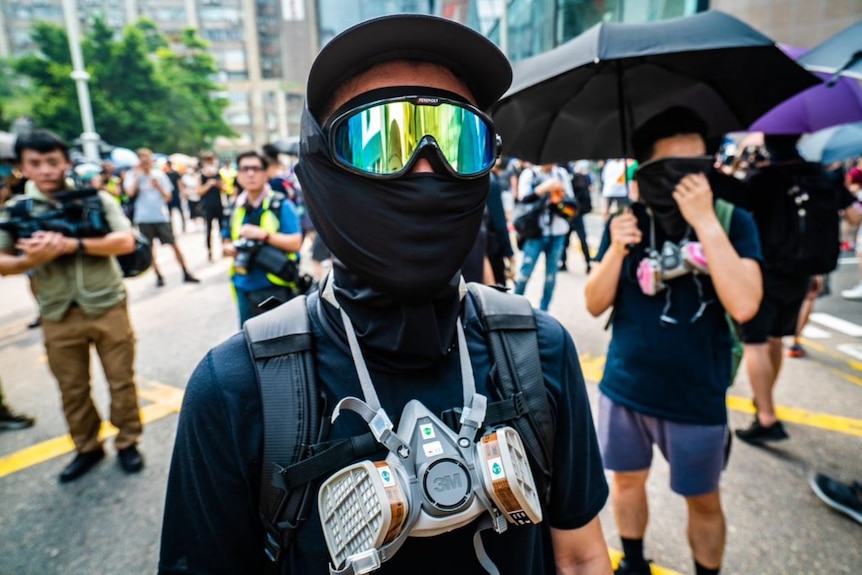 A person wearing a black shirt, googles and black fabric covering their mouth. Around their neck is a gas mask.