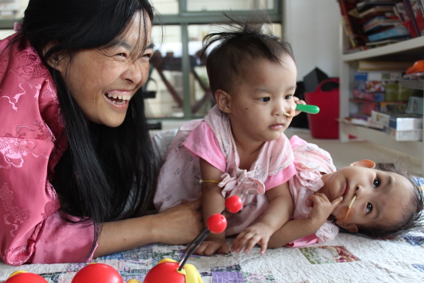 Conjoined twin toddler girls, both wearing pink, lay on the floor in a playroom with their smiling mother lying next to them