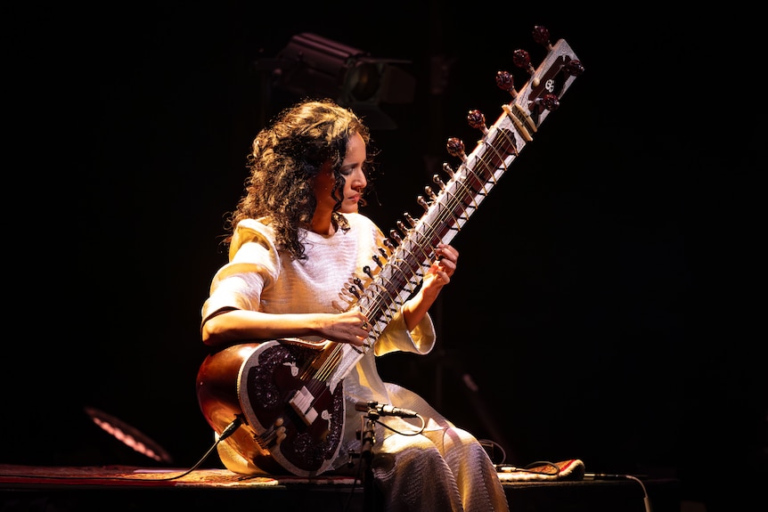 A woman sits on stage against a black background, playing the sitar. 