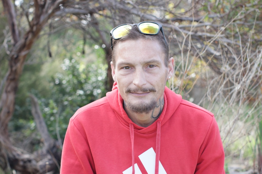 A man with a red hoodie and sunglasses on the top of his head looks at the camera.