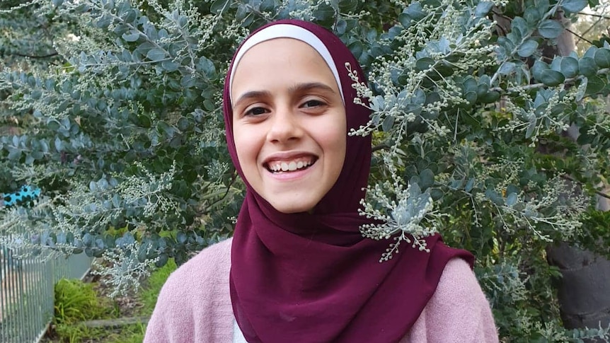 A smiling teen wearing a hijab in front of greenery