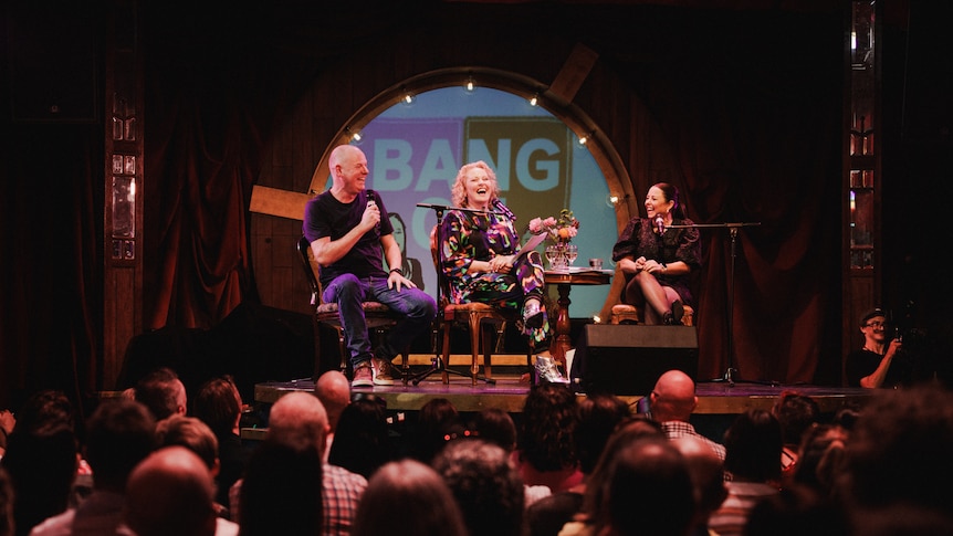 Tom Gleeson, Zan Rowe and Myf Warhurst are seated on a stage in front of an audience laughing